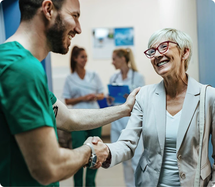 Smiling doctor in green scrubs shaking hands with an elderly female patient in a clinic hallway