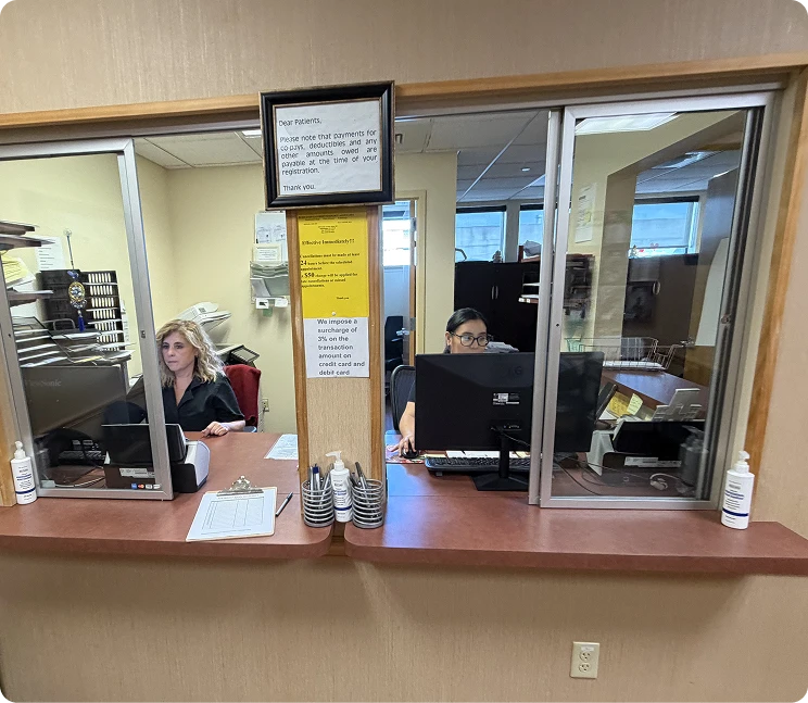 Medical office reception desk with two staff members working behind a sliding glass window partition